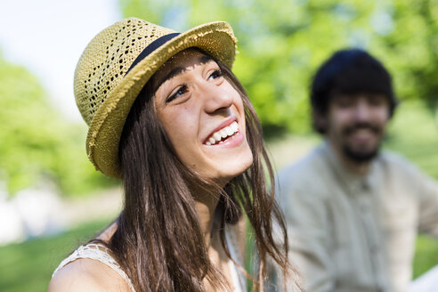 Porträt einer glücklichen jungen Frau in einem Park mit ihrem Freund im Hintergrund - GIOF04693