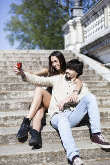 Glückliches junges Paar sitzt auf einer Treppe und macht ein Selfie mit seinem Smartphone - GIOF04698
