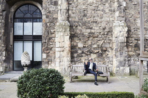 UK, London, älterer Geschäftsmann sitzt auf einer Bank in einem Innenhof und entspannt sich, während er mit Kopfhörern Musik hört, lizenzfreies Stockfoto
