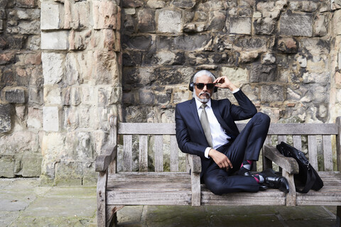 Porträt eines gut gekleideten älteren Geschäftsmannes mit Sonnenbrille und Kopfhörern, der auf einer Bank sitzt, lizenzfreies Stockfoto