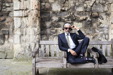 Porträt eines gut gekleideten älteren Geschäftsmannes mit Sonnenbrille und Kopfhörern, der auf einer Bank sitzt - IGGF00614