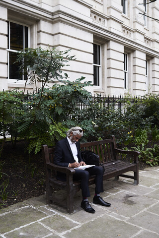 Älterer Geschäftsmann sitzt auf einer Bank im Freien und schreibt Notizen in ein Notizbuch, lizenzfreies Stockfoto
