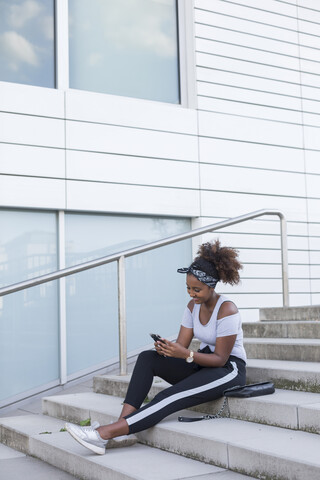 Lächelnde junge Frau sitzt auf einer Treppe und schaut auf ihr Smartphone, lizenzfreies Stockfoto