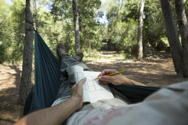 Man lying in hammock in the woods relaxing with puzzle book, partial view - JPTF00010