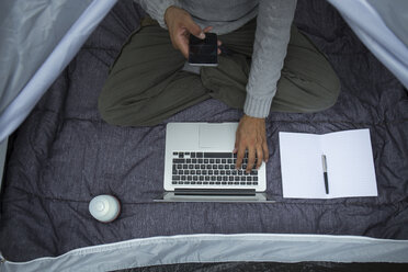 Mann sitzt im Zelt und benutzt Laptop und Mobiltelefon, Teilansicht - JPTF00016
