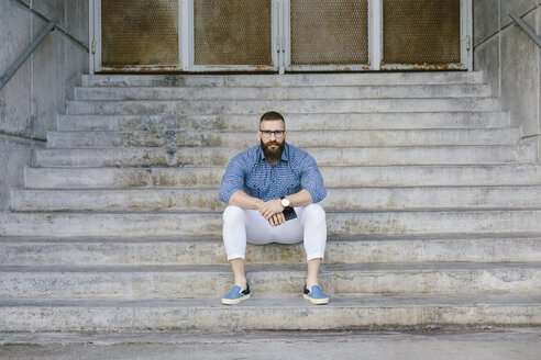 Porträt eines bärtigen Hipster-Geschäftsmannes mit Smartphone auf einer Treppe sitzend - FMGF00014
