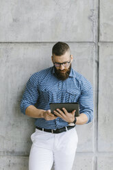 Bärtiger Hipster-Geschäftsmann mit Brille und kariertem Hemd lehnt an der Wand und benutzt ein Tablet - FMGF00022