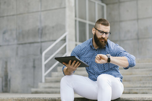 Porträt eines bärtigen Hipster-Geschäftsmannes, der mit einem digitalen Tablet auf einer Treppe sitzt und die Zeit überprüft - FMGF00024