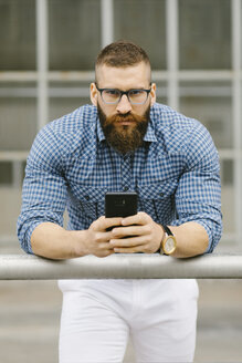 Porträt eines bärtigen Hipster-Geschäftsmannes mit Smartphone, der sich auf ein Geländer stützt - FMGF00025