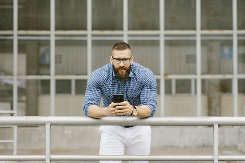 Porträt eines bärtigen Hipster-Geschäftsmannes mit Smartphone, der sich auf ein Geländer stützt - FMGF00026
