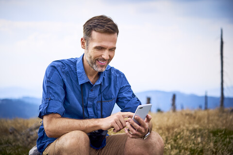 Lächelnder Mann, der sich beim Wandern ausruht und sein Mobiltelefon überprüft, lizenzfreies Stockfoto