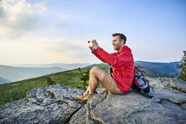 Man sitting on rock taking picture with his cell phone during hiking trip in the mountains - BSZF00731