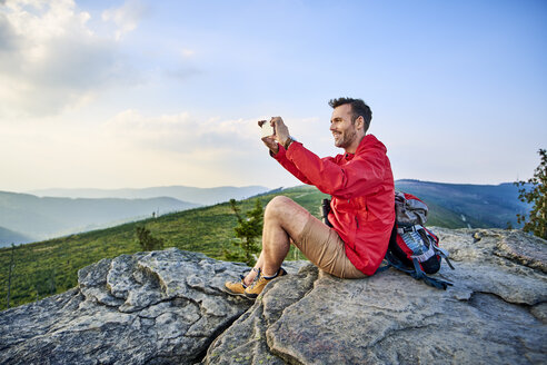 Mann sitzt auf einem Felsen und fotografiert mit seinem Handy beim Wandern in den Bergen - BSZF00731