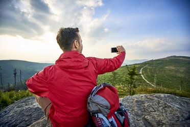 Man sitting on rock taking picture with his cell phone during hiking trip in the mountains - BSZF00732