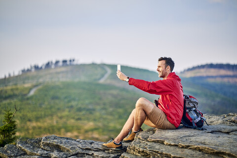 Mann sitzt auf einem Felsen und macht ein Selfie mit seinem Handy beim Wandern in den Bergen, lizenzfreies Stockfoto