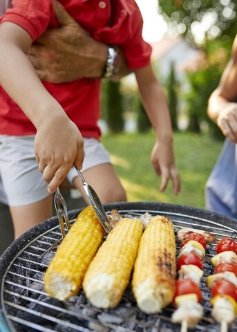 Junge dreht einen Maiskolben beim Grillen im Garten, lizenzfreies Stockfoto