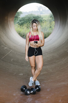 Sportliche Frau steht in einer Röhre mit Hanteln und Kopfhörern und schaut auf ihr Handy - FMGF00040