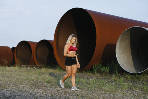 Sportliche Frau mit Handy und Kopfhörern beim Spaziergang in einem Industriegebiet, lizenzfreies Stockfoto