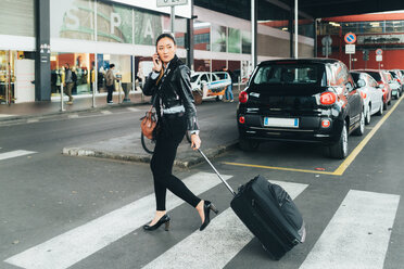 Woman walking outdoors, pulling wheeled suitcase, talking on smartphone - CUF45178