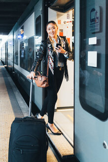 Frau steigt in einen Zug ein, zieht einen Rollkoffer, hält ein Smartphone - CUF45181