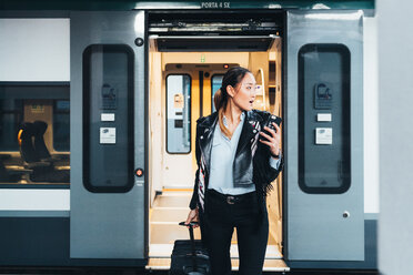 Frau steigt aus dem Zug, zieht einen Rollkoffer und hält ein Smartphone in der Hand - CUF45182