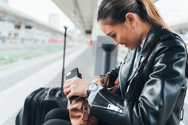 Woman sitting on train platform, looking through handbag, suitcase beside her, holding smartphone - CUF45183