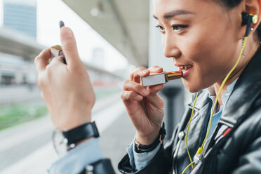 Woman sitting on train platform, applying lipstick, wearing earphones - CUF45185