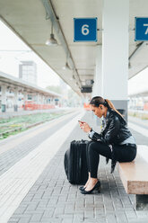 Woman sitting on train platform, using smartphone, suitcase beside her - CUF45186