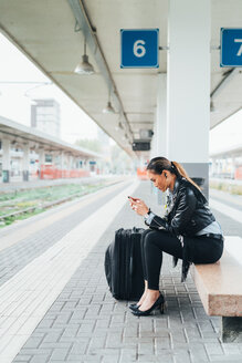 Frau sitzt auf dem Bahnsteig und benutzt ihr Smartphone, Koffer neben ihr - CUF45186