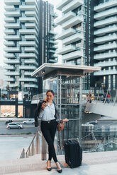 Businesswoman, outdoors, holding wheeled suitcase, using smartphone - CUF45196