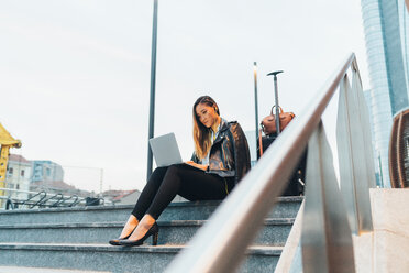 Businesswoman, sitting outdoors on steps, using laptop, luggage beside her, low angle view - CUF45200