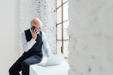 Businessman sitting on windowsill using laptop and smartphone - CUF45271