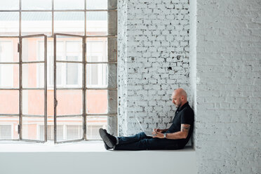 Man sitting on windowsill using laptop, full length, side view - CUF45286