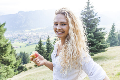 Deutschland, Bayern, Oberammergau, Porträt einer lächelnden jungen Frau, die auf einer Bergwiese einen Apfel isst - TCF05848