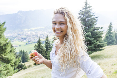 Germany, Bavaria, Oberammergau, portrait of smiling young woman eating an apple on mountain meadow - TCF05848