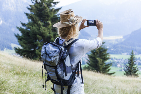 Deutschland, Bayern, Oberammergau, junge Frau wandert und macht ein Handyfoto auf einer Bergwiese, lizenzfreies Stockfoto