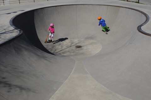 Brother and sister on scooters playing at sunny skate park stock photo