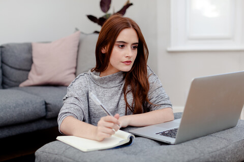 Frau schreibt Notizen in Notizblock von Laptop, lizenzfreies Stockfoto