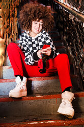 Junge Frau sitzt auf einer Treppe, benutzt ein Smartphone und lächelt - CUF45716