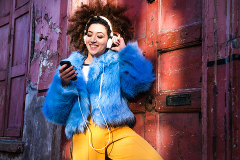 Porträt einer Frau mit Afrohaar und Pelzmantel, die über Kopfhörer auf einem Smartphone Musik hört, lizenzfreies Stockfoto