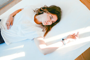 Young woman relaxing on bed, arm resting in ray of light, thoughtful expression - CUF46043