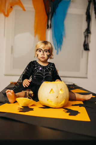 Porträt eines kleinen Mädchens mit bemaltem Gesicht und Kostüm auf einem Tisch mit Jack O'Lantern sitzend, lizenzfreies Stockfoto