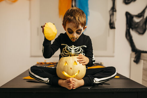Porträt eines kleinen Jungen mit bemaltem Gesicht und Kostüm, der auf einem Tisch sitzt und in eine Jack O'Lantern schaut - JRFF01881