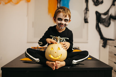 Porträt eines lächelnden kleinen Jungen mit bemaltem Gesicht und Kostüm, der auf einem Tisch mit Jack O'Lantern sitzt - JRFF01882