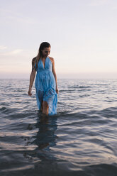 Young woman wearing blue dress walking in water at seashore by sunset - MAUF01720