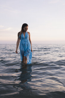 Junge Frau in blauem Kleid geht bei Sonnenuntergang im Wasser am Meer spazieren - MAUF01720