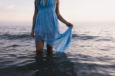 Young tattooed woman wearing blue dress standing in the sea by sunset, partial view - MAUF01721