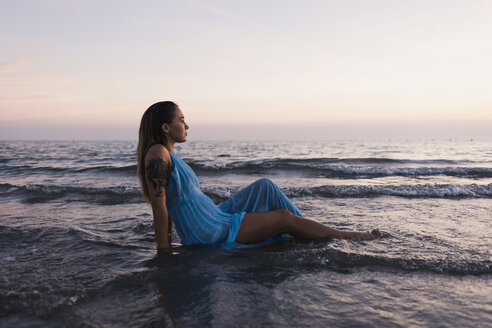 Junge tätowierte Frau in blauem Kleid sitzt im Wasser am Meeresufer und beobachtet den Sonnenuntergang - MAUF01722