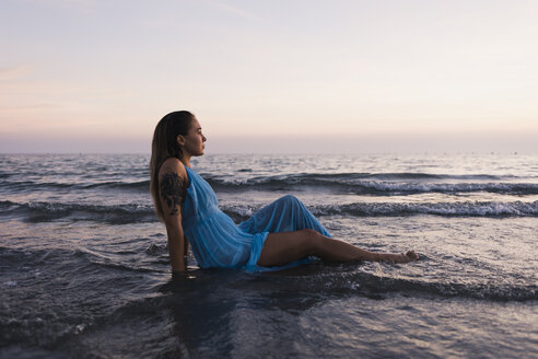 Young tattooed woman wearing blue dress sitting in water at seashore watching sunset - MAUF01722