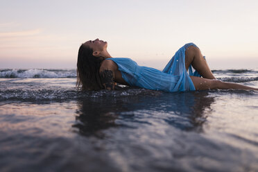Young tattooed woman wearing blue dress lying in water at seashore by sunset - MAUF01723